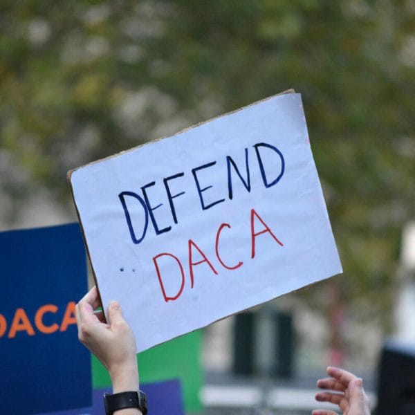 A hand holding a small white sign at a rally that says "Defend DACA"