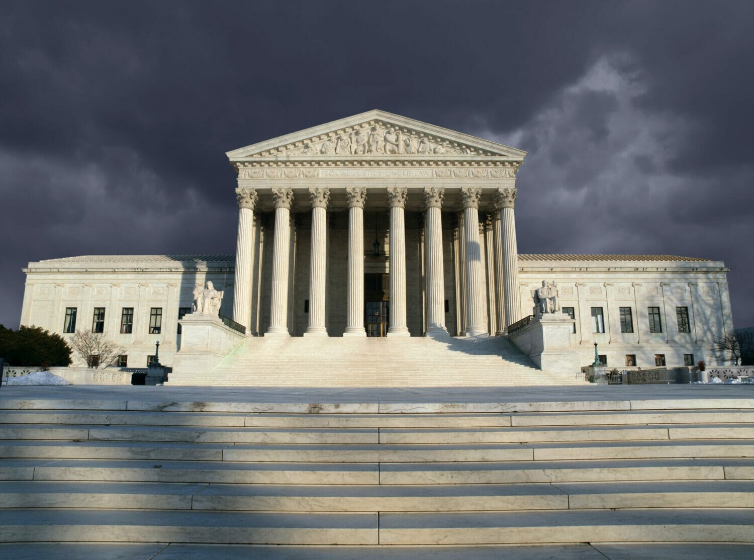 Straight-on view of the U.S. Supreme Court, somewhat lit in the foreground with ominous storm clouds gathering behind.