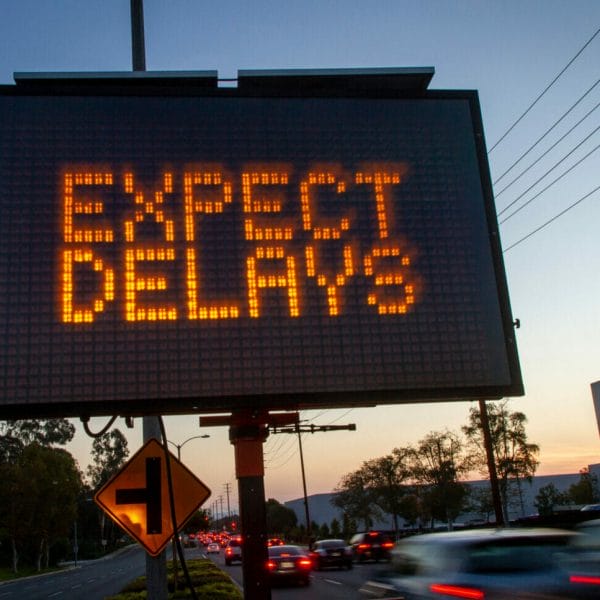 Lighted traffic sign reading "expect delays" in capital organe letters, with a dusk sky in the background and cars driving by in the foreground.