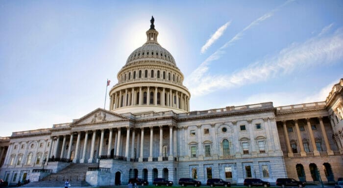 The U.S. Capitol building backlit by morning sunlight, with a blue sky and light, thin lines of clouds emanating from behind the rotunda.