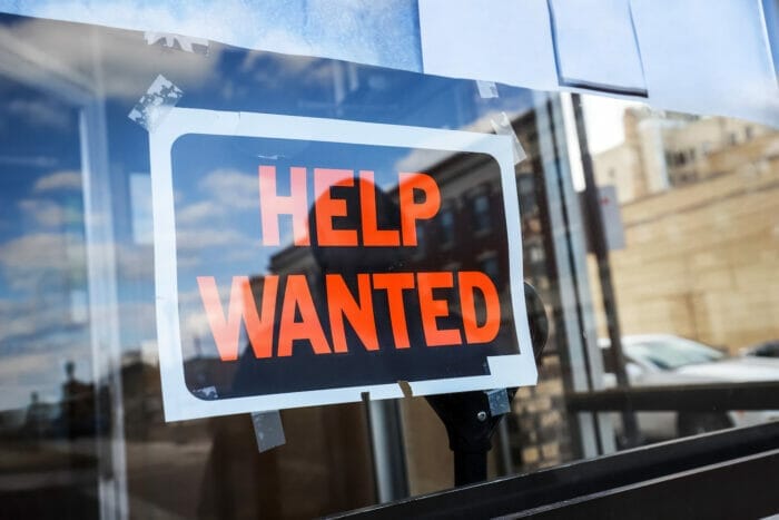 Reflection of a man looking at a black and orange help wanted sign in a business window