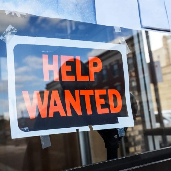 Reflection of a man looking at a black and orange help wanted sign in a business window