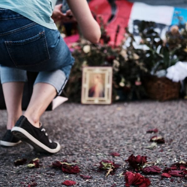 A woman in denim capris and black and white sneakers visits a memorial at the site of the shooting at Walmart in El Paso Texas. Out of focus beyond the woman are flowers, signs, and framed photos.
